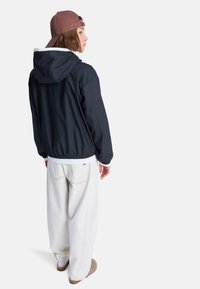 Ναυτικό μπλε αμάνικο μπουφάν με κουκούλα, λευκή επένδυση και ελαστικό τελείωμα, συνδυασμένο με άνετο λευκό παντελόνι και καφέ παπούτσια χωρίς κορδόνια. Καθημερινή εμφάνιση από πίσω.