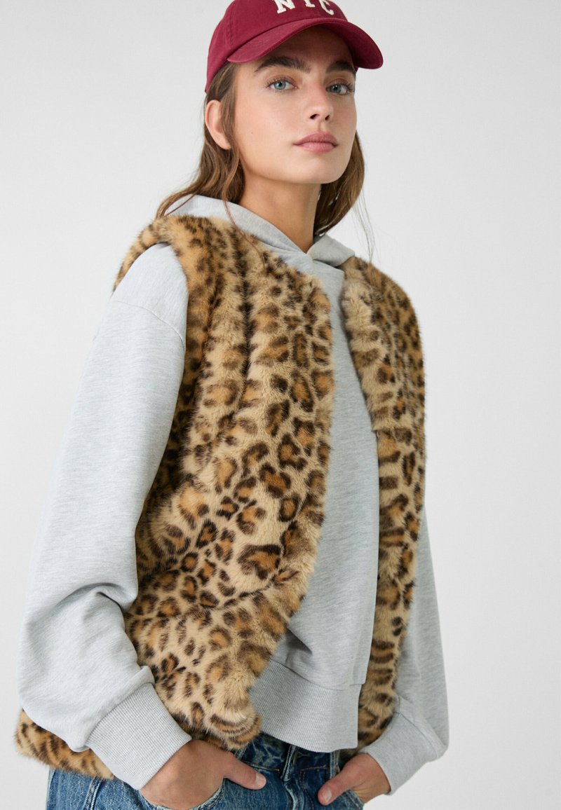 Gilet in pelliccia sintetica con stampa leopardata indossato sopra una felpa con cappuccio grigia chiara. Il gilet presenta una texture morbida, un design aperto nella parte anteriore e nessun fissaggio visibile.