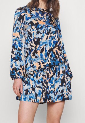Femme portant une mini-robe à manches longues, coupe ample, avec un motif abstrait bleu, noir et pêche sur un fond uni.