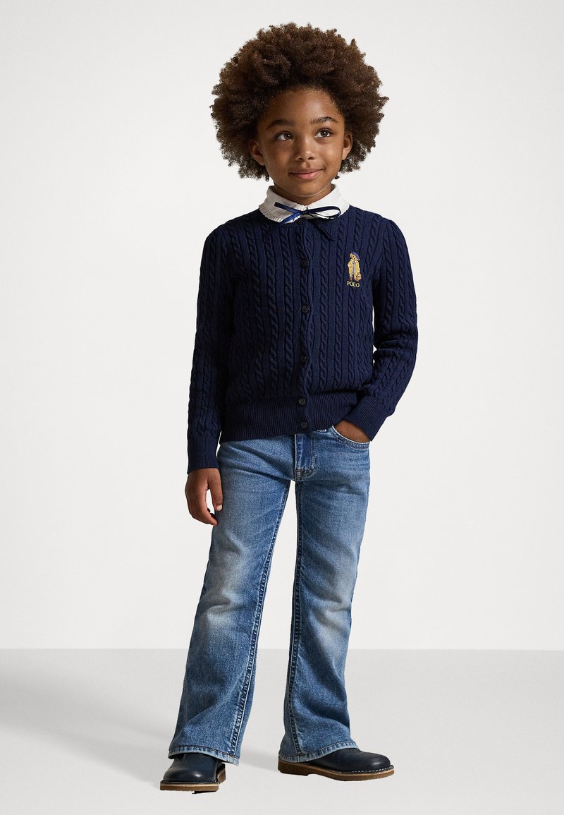 Marinblå stickad cardigan med flätmönster, knappstängning och broderad logotyp. Tillsammans med blekta blå utsvängda jeans och svarta skor. Vit skjorta med krage under.