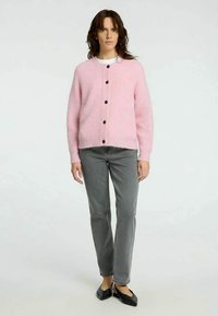 Mulher de pé, vestindo um cardigan rosa com botões, uma blusa branca, jeans cinzentos e sapatilhas pretas, contra um fundo claro e liso.