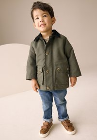 Veste vert olive avec un col noir, des fermetures à boutons et deux poches avant, associée à un jean bleu retroussé au niveau des chevilles et des baskets marron.