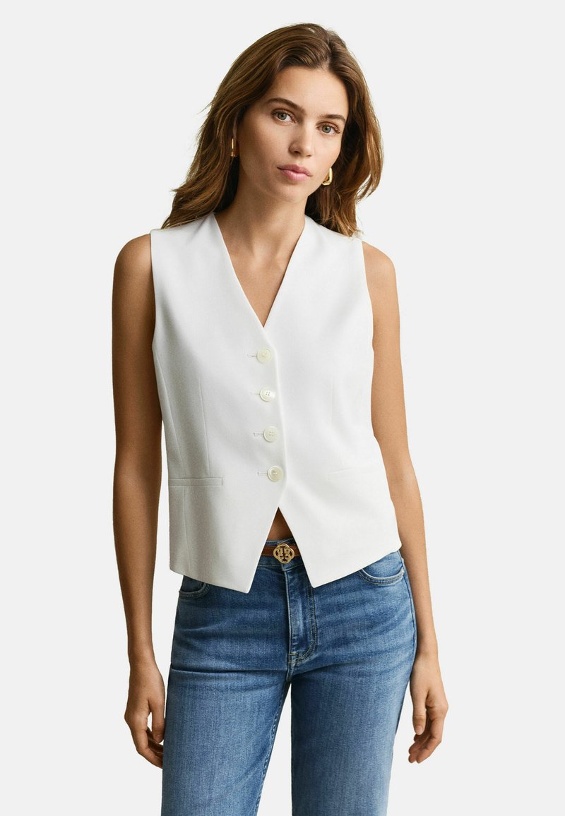 Femme portant un gilet blanc sans manches à boutons et un jean bleu, debout les bras détendus contre un fond clair uni.