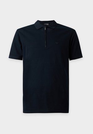 Jeune homme portant des lunettes de soleil noires, un polo noir à manches courtes, un pantalon gris et des chaussures habillées noires, debout devant un fond blanc uni.
