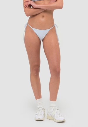 Kobieta ubrana w jasnoniebieskie bikini z wiązaniami po bokach, białe skarpetki do kostek oraz białe buty sportowe, stoi z założonymi rękami na tle jednolitego koloru.