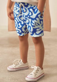 Shorts à motifs bleus et blancs avec une taille à cordon, comportant des poches latérales. Associés à des sneakers en toile blanches avec des accents rouges.