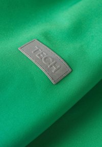 Tissu vert avec une étiquette rectangulaire grise cousue, embossée du mot "TECH" en lettres en relief.