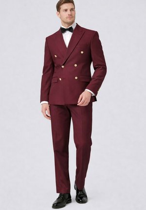 Homme portant un costume croisé bordeaux avec des boutons dorés, une chemise blanche, un nœud papillon noir et des chaussures de ville noires, debout sur un fond uni.