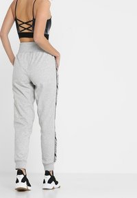 Grijze trainingsbroek met elastische tailleband, zijstrepen en opgerolde enkels, gecombineerd met zwart-witte sneakers. Het achteraanzicht toont een kruislingse ontwerp aan de bovenkant.