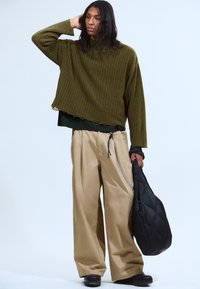 Personne avec de longs cheveux bruns portant un pull côtelé vert olive, un pantalon beige à larges jambes avec un accessoire en chaîne, tenant un sac matelassé noir.