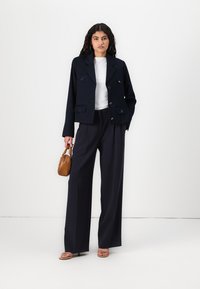 Blazer marine avec deux poches avant, boutons croisés, associé à un pantalon large taille haute et un haut léger. Petit sac à main marron.