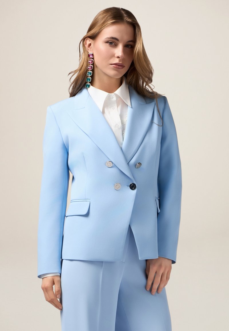 Blazer doppiopetto azzurro chiaro con bottoni argentati, abbinato a una camicia bianca con colletto. Presenta un tessuto liscio e una vestibilità su misura.