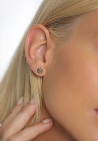 Guld ørestikker med en tekstureret overflade, hvilende på en models øre. Designet er rundt, med en diskret glans og små dimensioner.