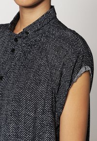 Chemise noire à manches courtes avec un motif tacheté blanc, dotée d'un col boutonné et de manches roulées, fabriquée en tissu léger.