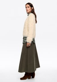 Sweter z sztucznego futra w kolorze kremowym, zielona koszula w kratę nałożona pod spód, oliwkowa maxi spódnica, brązowe botki na obcasie, z pierścionkami na rękach.