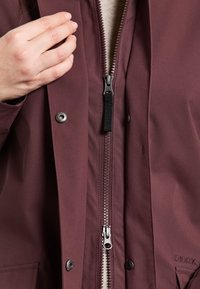 Bordeaux waterproof jack met een rits aan de voorkant, drukknoopjes en een gladde afwerking. Zwarte ritsenstrop en subtiele branding op de stof.