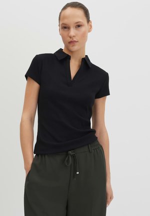 Femme portant une chemise noire à manches courtes avec col et un pantalon vert foncé à cordon, debout devant un fond clair uni.