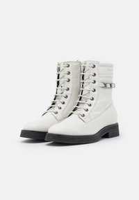 Bottines blanches en cuir avec un laçage à l'avant, bout rond et semelle en caoutchouc texturé noir. Présente une sangle décorative et une fermeture éclair sur le côté.