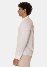 Camicia bianca a maniche lunghe realizzata in un tessuto leggero, con colletto appuntito e polsini con bottoni. Design semplice e privo di decorazioni.