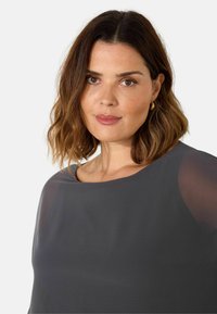 Plus-size blůza v tmavě šedé barvě s kulatým výstřihem. Má průhledné rukávy a hladkou texturu, což doplňuje elegantní design.