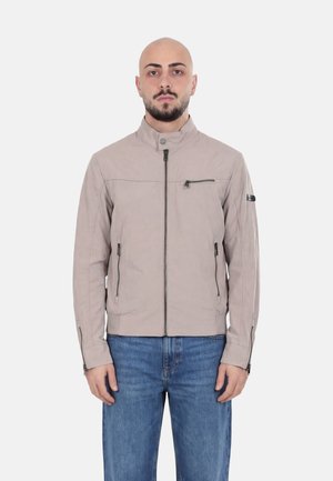 Uomo calvo con barba indossa una giacca beige chiaro con cerniere e tasche con cerniere e jeans blu, in piedi davanti a uno sfondo chiaro uniforme.