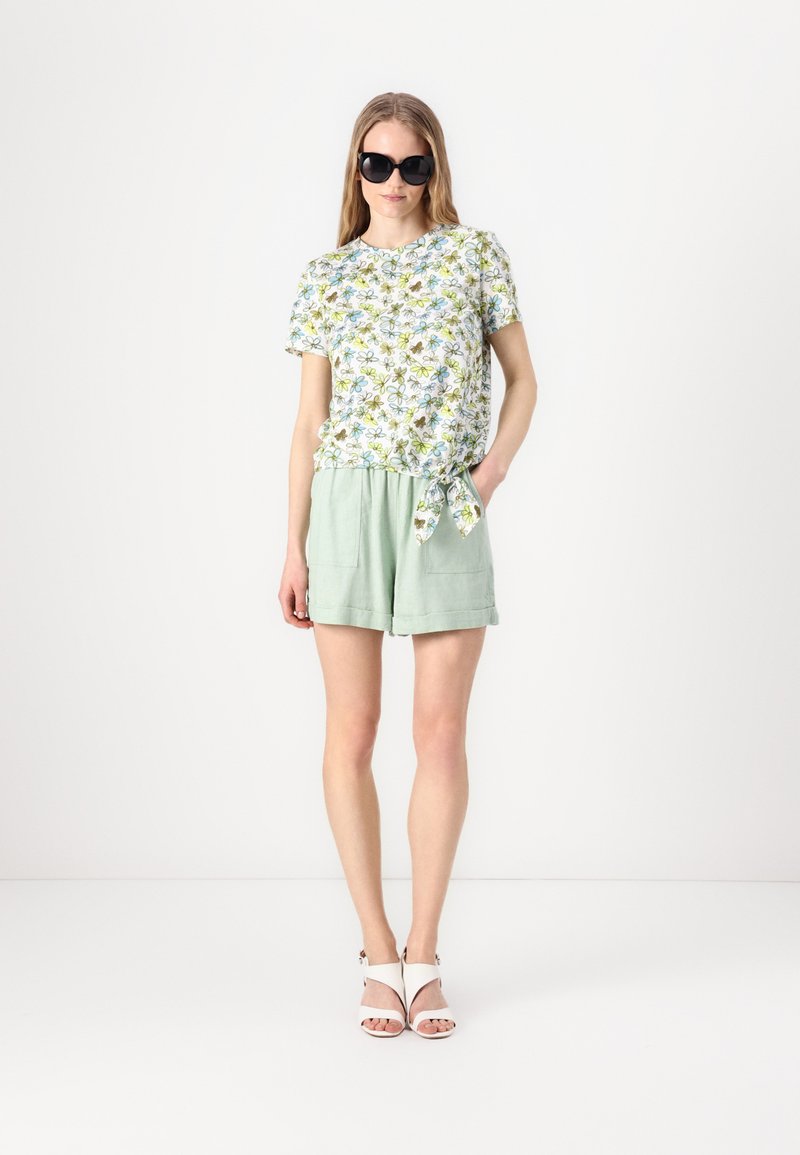Camicia a maniche corte con motivo floreale e nodo frontale, abbinata a pantaloni corti a vita alta verde chiaro e sandali bianchi con cinturino.