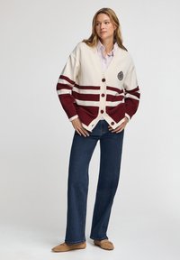 Krämfärgad cardigan med vinröda ränder, knappstängning och broderad emblem; kombinerad med blå jeans och beige slip-on skor. Slät textur.