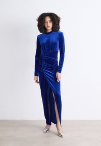 Vestido de veludo azul royal com mangas longas, apresentando franzido lateral e uma fenda até à coxa. Combinado com sapatos de salto metálicos.