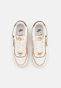 Les baskets Nike Air Force 1 présentent une tige en cuir blanc avec des accents orange et marron, des superpositions texturées et des perforations bien marquées sur la boîte à orteils.