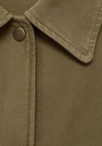 Chemise à col olive en tissu texturé, avec une fermeture à boutons-pression et des détails de couture visibles près du col et de la patte de boutonnage.