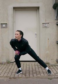 Kvinde i sort sportstøj strækker benene på et stenbelagt fortov foran en lukket beige dør på en betonvæg.
