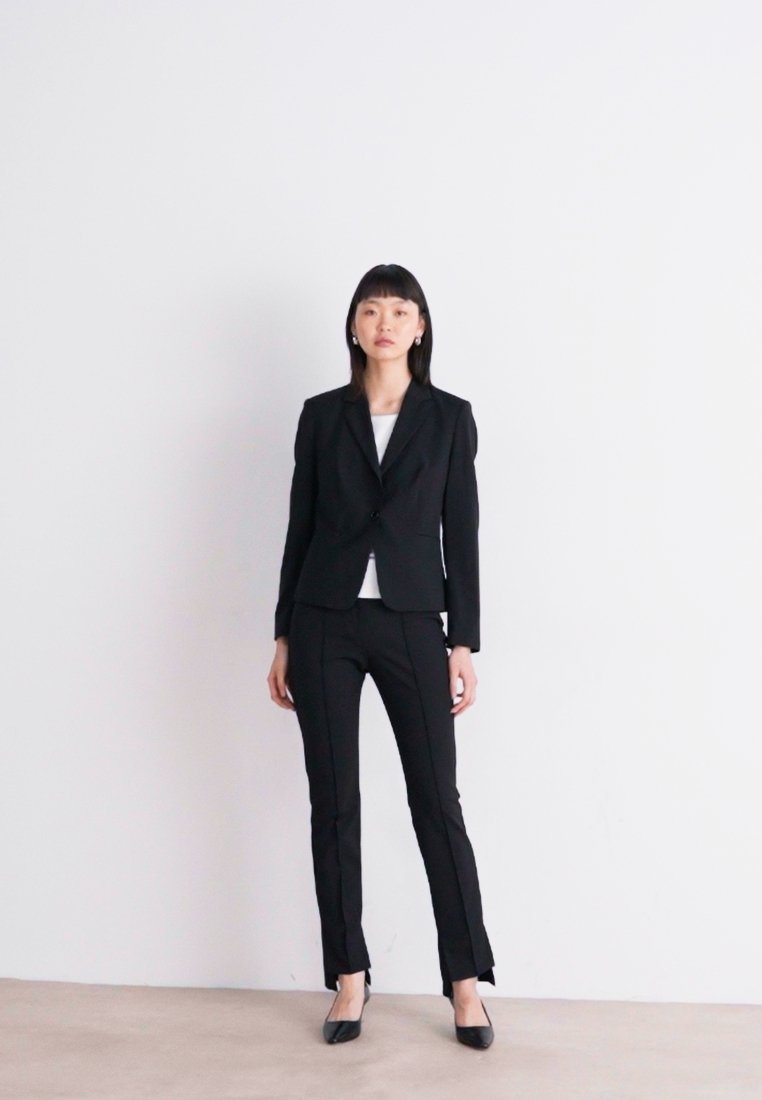Costume noir ajusté avec blazer structuré et pantalon slim, associé à un haut blanc. Tissu lisse, design minimaliste et coupe classique.