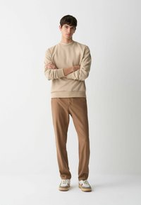 Mlad moški stoji z rokami prekrižanimi, oblečen v bež pulover, rjave hlače in bele teniske na enostavni beli podlagi.