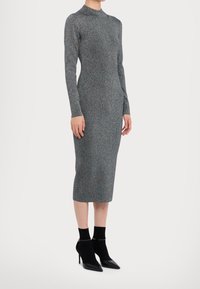 Robe ajustée à manches longues en tissu gris texturé avec un léger éclat. Longueur mi-mollet, encolure ronde, associée à des escarpins noirs à talons pointus.