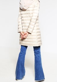 Béžová péřová bunda s horizontálním prošíváním, kapucí a dlouhými rukávy, v kombinaci s modrými zvonovými džíny a neutrálními botami.