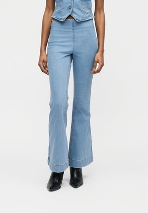 Femeie purtând blugi evazați cu talie înaltă, de culoare albastru deschis, vestă din denim scurtă și cizme negre cu toc mediu, pe fundal alb simplu.