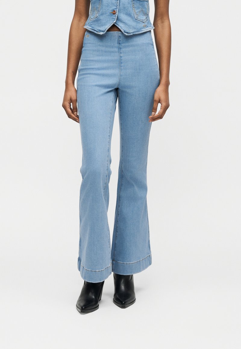 Γυναίκα με ανοικτό μπλε ψηλόμεσα τζιν καμπάνα, κοντό γιλέκο από τζιν και μαύρα μποτάκια με τακούνι, σε απλό λευκό φόντο.