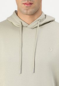 Hoodie bege claro com cordão, equipado com terminais prateados. Apresenta um logótipo embutido subtil no peito. Textura suave.