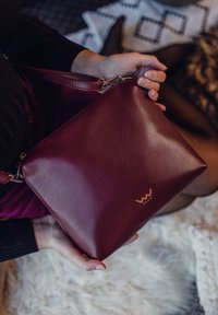 Burgundská kožená kabelka s hladkým povrchem, se zipovým uzávěrem a zlatým logem. Obsahuje popruh pro nošení v ruce nebo na rameni.
