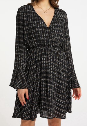 Kobieta ubrana w czarną sukienkę w wzory z długimi rozszerzanymi rękawami, dekoltem w kształcie litery V i marszczonym detalem w talii.