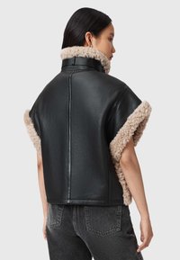 Moteris dėvi trumpomis rankovėmis juodą odinę striukę su smėlio spalvos avies vilnos apdaila apykaklėje ir rankogaliuose, derančią su tamsiai pilkais džinsais.