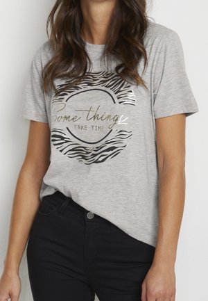 Femme portant un t-shirt gris clair avec un cercle à motif zèbre et le texte « Certaines choses prennent du temps », assorti à un pantalon noir.