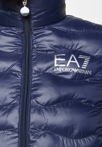 Marinblå puffy-jacka med kviltad textur, framzipperstängning och EA7-logotyp i silver. Har en h�ög krage för extra värme.