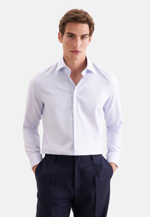 Jeune homme portant une chemise bleu clair rentrée dans un pantalon bleu marine foncé, les mains dans les poches, sur un fond uni.