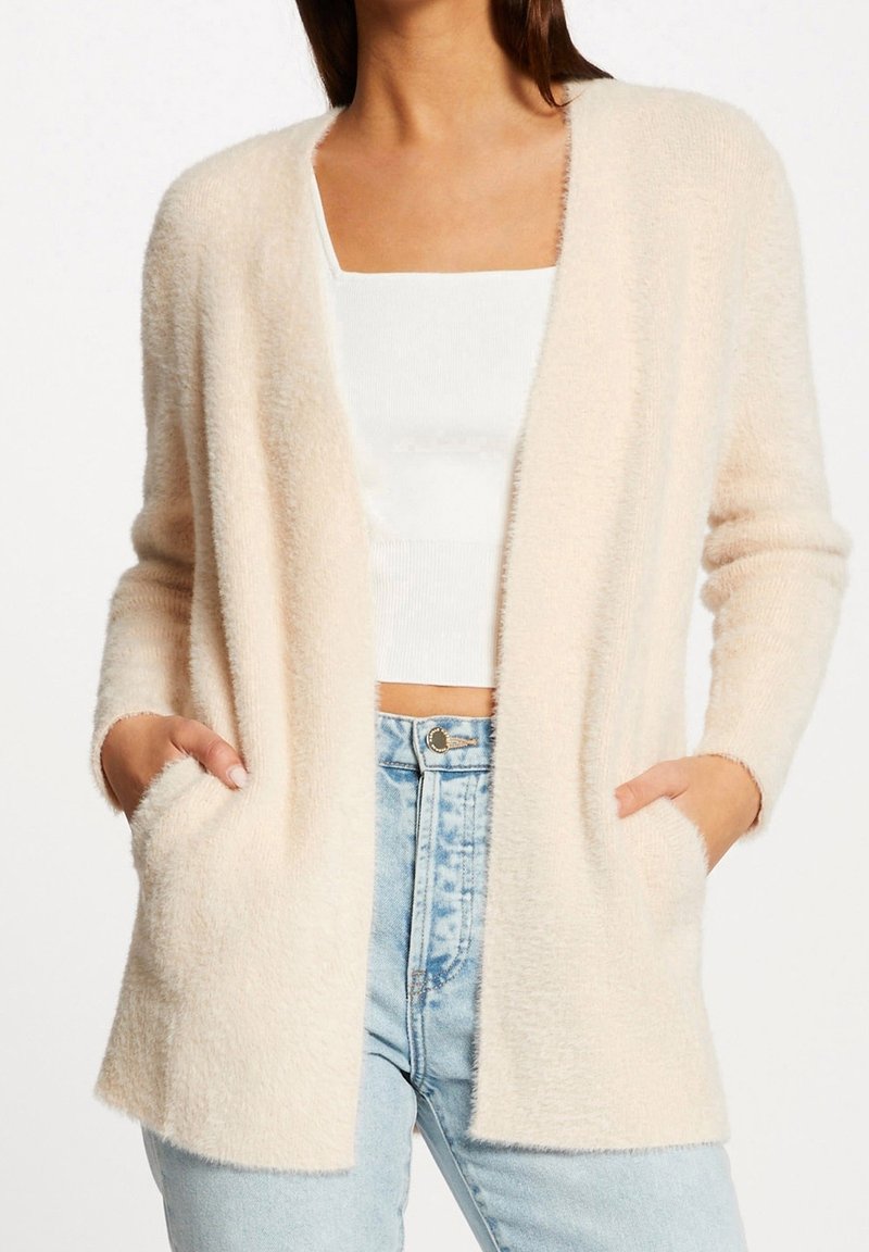 Femme portant un cardigan crème pelucheux ouvert sur un crop top blanc et un jean taille haute bleu clair, les mains dans les poches.