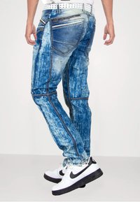 Verbleekte blauwe denimjeans met lichte en donkere wasspatronen, voorzien van meerdere zakken en contrasterende stiksels. Draag ze met witte sneakers.