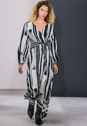 Žena sa kovrčavom plavom kosom u crnoj, bijeloj i sivoj prugastoj zavojnoj haljini s dugim rukavima i crnim čizmama s špicastim vrhovima hoda u zatvorenom prostoru.