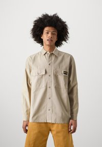 Beige long-sleeve shirt with button-down front, two chest pockets, and black label, paired with yellow shorts, on a neutral background.