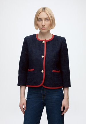 Femme aux cheveux blonds courts portant une veste bleu marine texturée avec un bord rouge et des boutons dorés, associée à un jean bleu, debout sur un fond blanc.