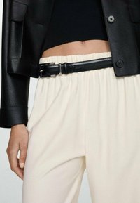 Schwarze Lederjacke, eng anliegendes schwarzes Oberteil und cremfarbene Hose mit elastischem Bund. Der schwarze Ledergürtel hat eine silberne Schnalle.